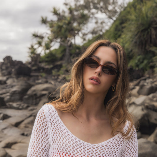 Woman wearing Alkira branded sunglasses and a white mesh top on a rocky beach