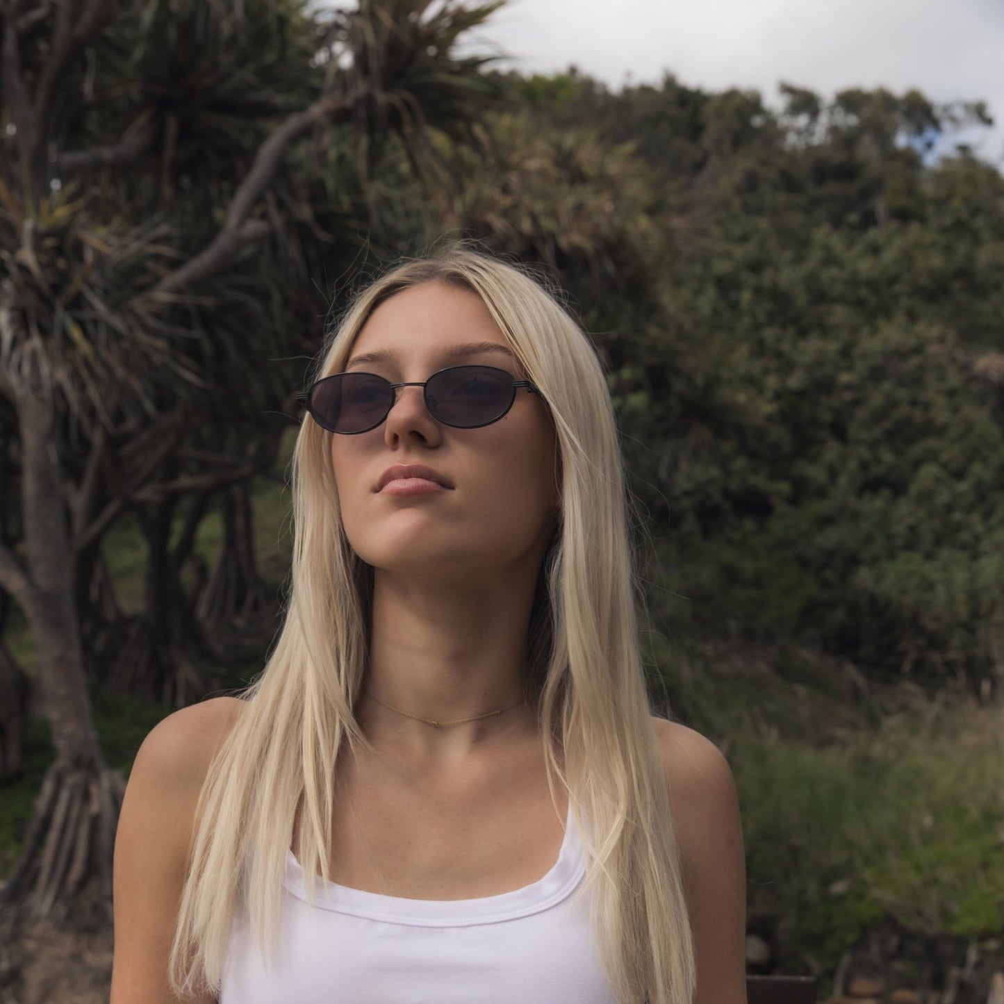 Woman wearing Alkira branded sunglasses and a white top outdoors with greenery in the background