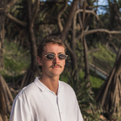Man wearing Alkira branded sunglasses and a white shirt in front of trees