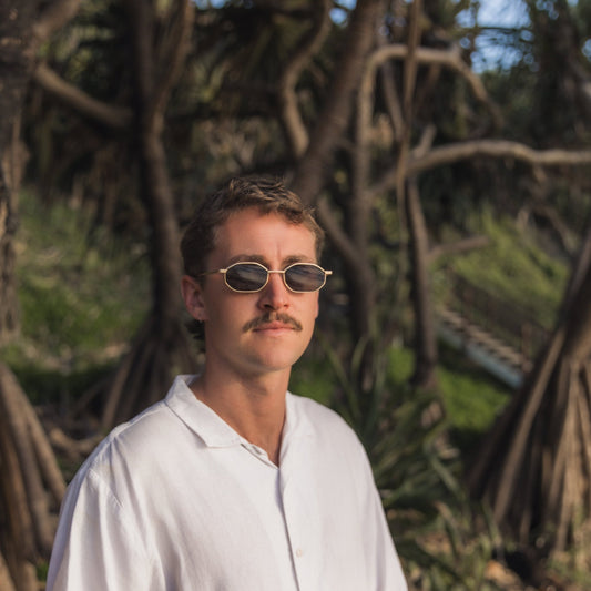 Man wearing Alkira branded sunglasses and a white shirt in front of trees