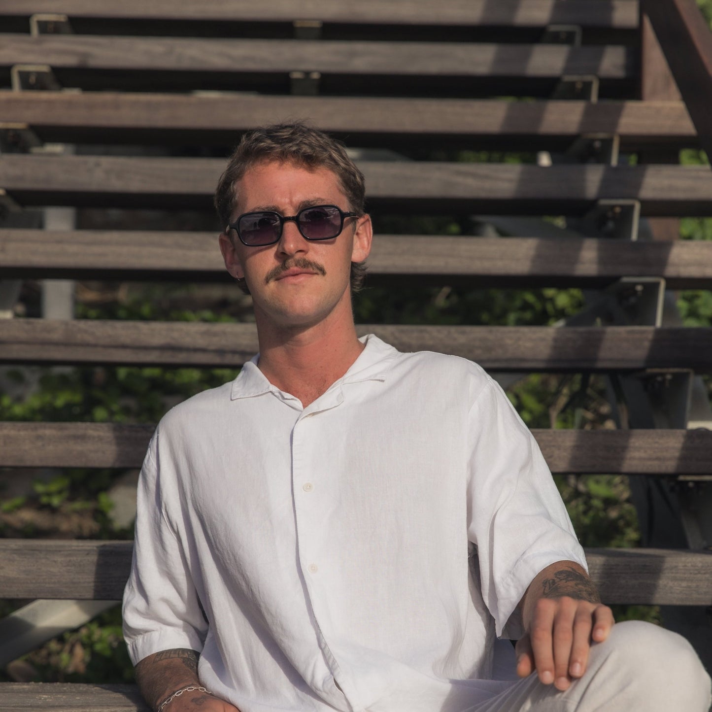 Man wearing Alkira branded sunglasses and a white shirt sitting on a wooden bench outdoors.