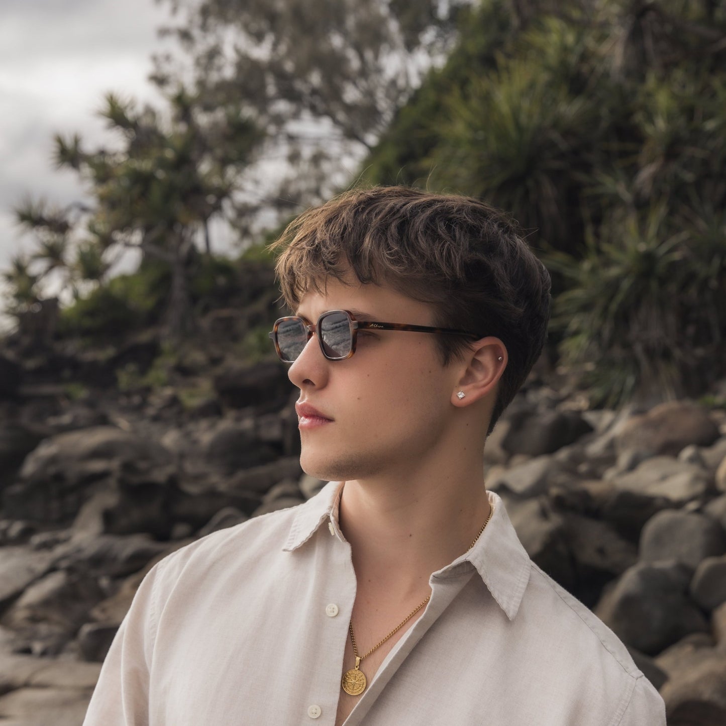 Person wearing Alkira branded sunglasses and a light shirt in a natural setting with rocks and greenery.