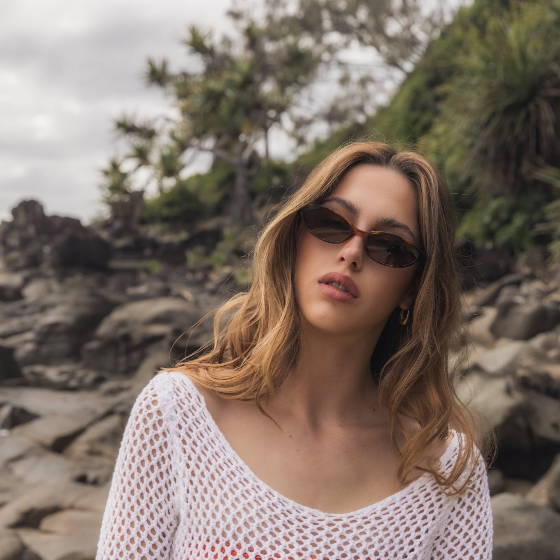 Woman wearing Alkira branded sunglasses and a white mesh top on a rocky beach