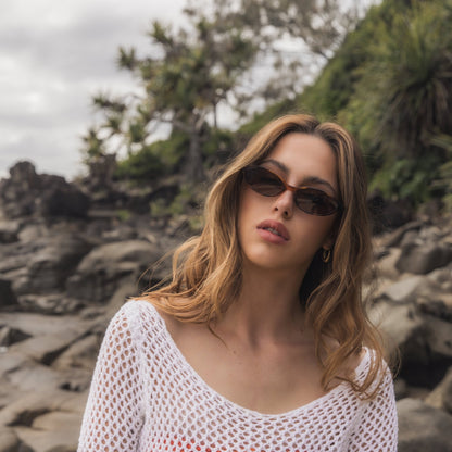 Woman wearing Alkira branded sunglasses and a white mesh top on a rocky beach