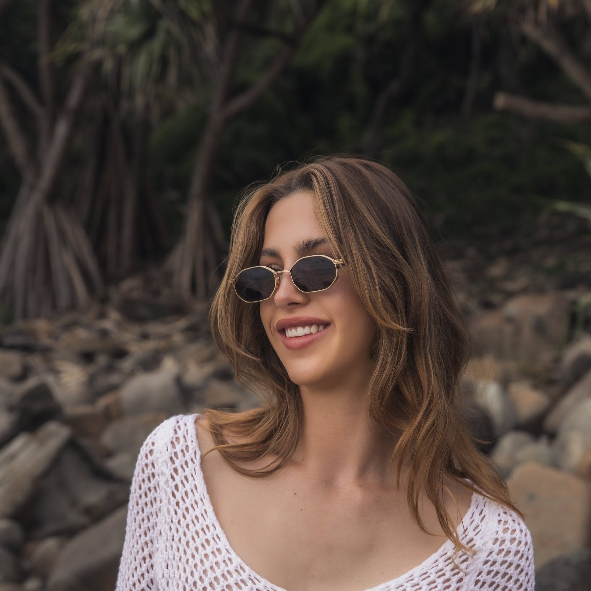 Woman wearing Alkira branded sunglasses and a white mesh top standing on a rocky beach.