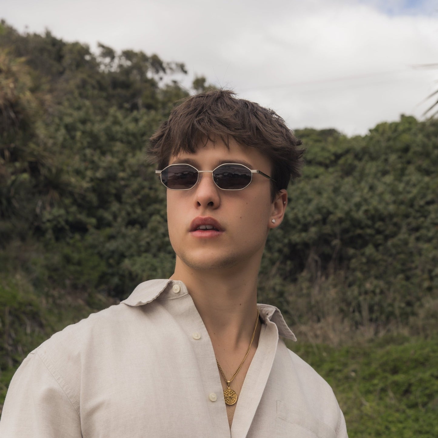 Person wearing Alkira branded sunglasses and a light-colored shirt outdoors with greenery in the background